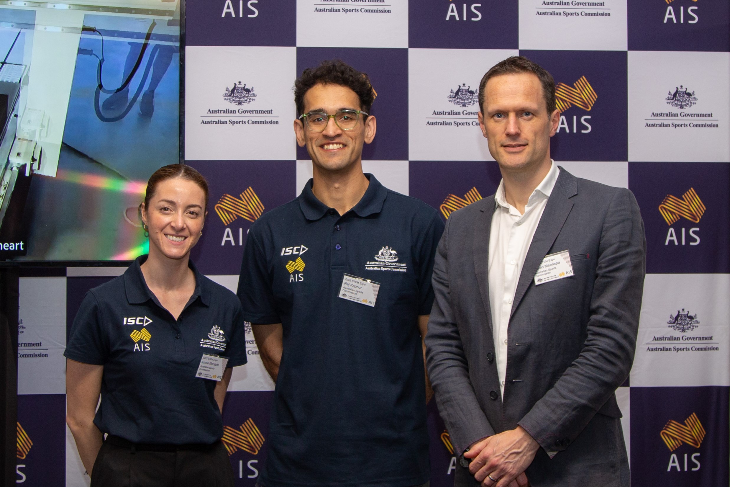 A photo of Senior Sports Dietitian Aimee Morabito, Sports Engineer Raj Kapoor and Chief Science Officer Paolo Menaspa standing in the AIS booth in the Great Hall in Parliament House